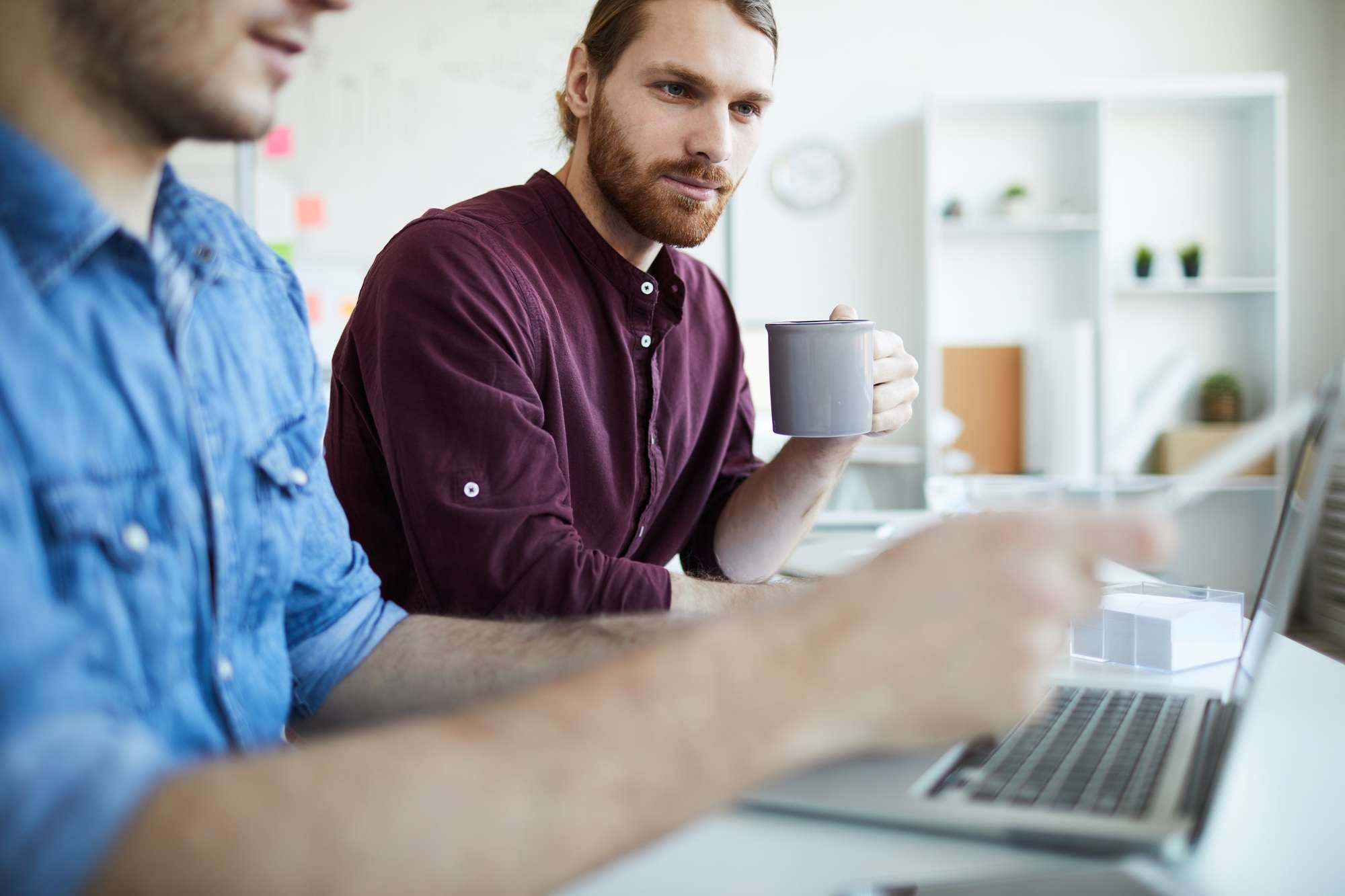Two men discussing e-commerce sales strategy while reviewing data on a laptop one holding a coffee mug in a modern office setting.