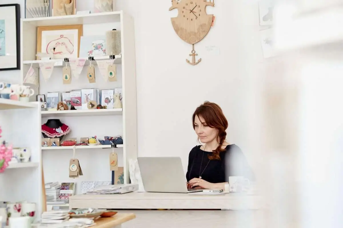 A woman sitting at a desk in a small gift shop, using a laptop.