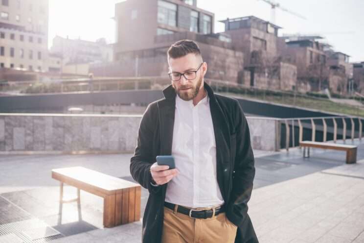 Young bearded man outdoor using smartphone texting searching online buying ecommerce